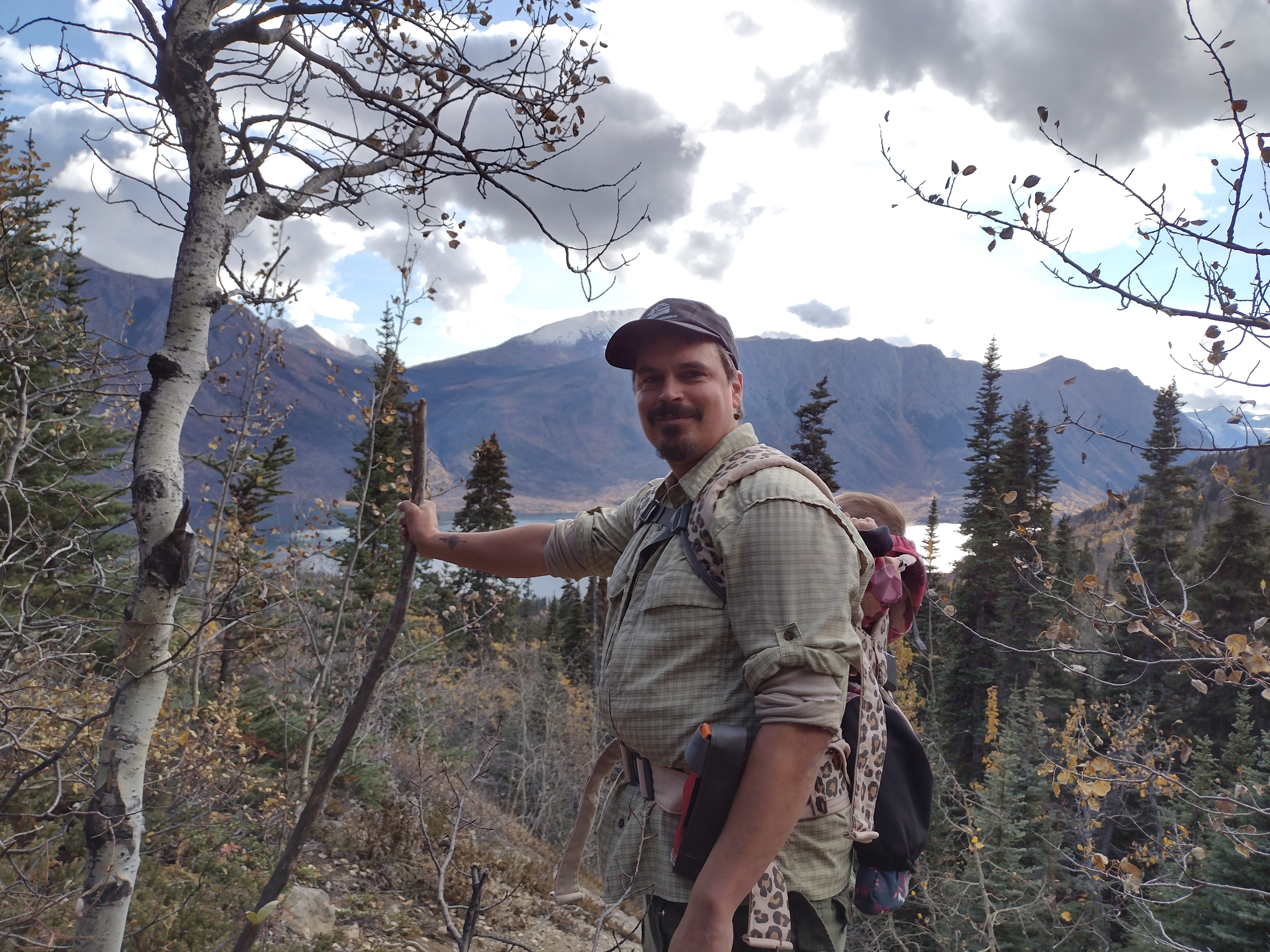 Chad Cote hiking with a baby carrier in the Yukon mountains, fall colours and a lake visible behind him